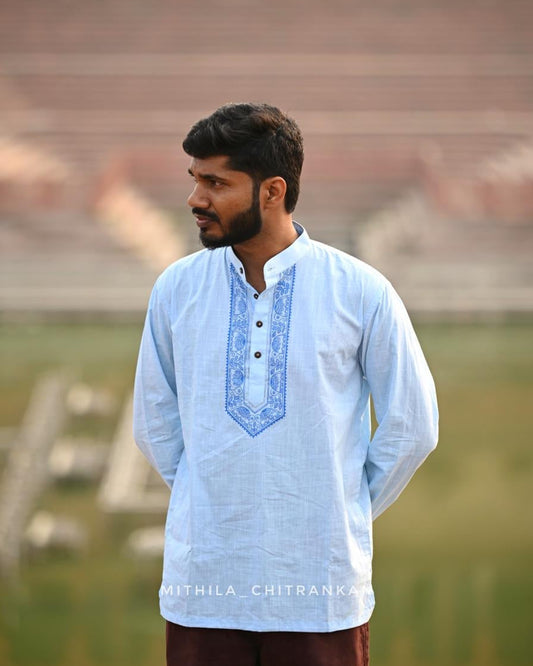 A man wearing a light blue long-sleeve kurta with blue embroidered detailing on the chest, standing in front of a blurred stadium background.